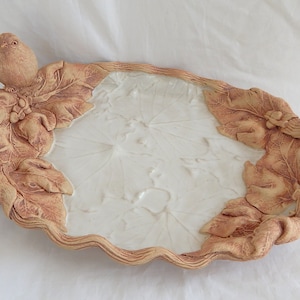 May include: A ceramic serving dish with a white glazed interior and a brown, textured exterior. The dish is shaped like a leaf and has a bird perched on the edge.