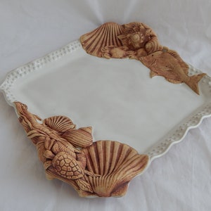 May include: White ceramic serving tray with a scalloped edge and raised brown clay sea life designs. The tray features a fish, seashells, and a sea turtle.