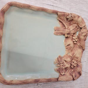Può includere: Vassoio da portata rettangolare in ceramica con interno azzurro e bordo marrone strutturato. Il lato destro presenta dettagli a rilievo di foglie, una libellula, farfalle e uva.