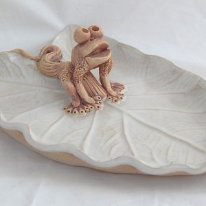 May include: A white ceramic dish shaped like a leaf with a brown ceramic frog sitting on top. The frog has large eyes and a wide mouth.