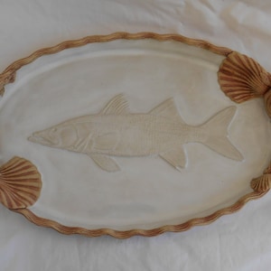 May include: White ceramic oval serving tray with a raised design of a fish and seashells around the edges. The tray has a brown glaze.