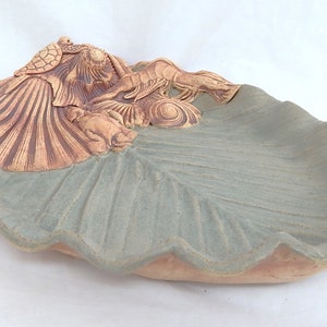 May include: A ceramic dish shaped like a seashell with a raised design of a turtle, a fish, and seaweed. The dish is glazed in a light green color with brown accents.