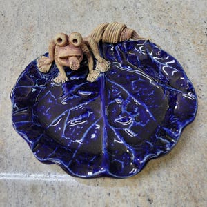 May include: A blue ceramic dish shaped like a leaf with a small ceramic frog on top. The frog has a brown and white glaze.