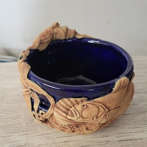 May include: A ceramic bowl with a deep blue interior and a tan exterior. The exterior is intricately carved with fish and other aquatic life. The bowl is likely handmade and suitable for decorative or functional use.