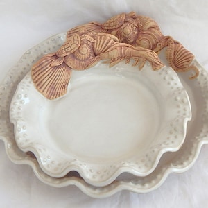 May include: A set of two white ceramic serving dishes with a scalloped edge and a raised dot pattern. The larger dish has a smaller dish nested inside. The dishes are decorated with a raised clay design of sea creatures, including a seahorse, a crab, and a turtle.