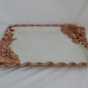 Puede incluir: Una bandeja de servir de cerámica con un interior esmaltado blanco y un exterior texturizado marrón. La bandeja presenta un borde festoneado y diseños esculpidos de vida marina, incluyendo peces y conchas.