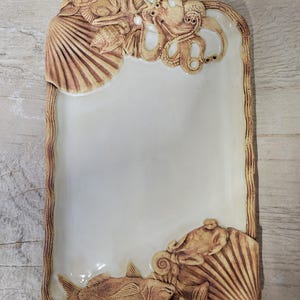 May include: Rectangular ceramic serving platter with a cream-colored center and a detailed brown border. The border features sculpted sea creatures, including a turtle, fish, octopus, and seashells, creating a coastal theme.