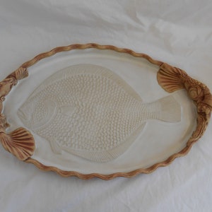 May include: A white ceramic serving tray with a raised design of a fish. The tray has a brown, scalloped edge and two brown, shell-shaped handles.