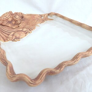 May include: A ceramic serving tray with a wavy, brown rim and a white interior. The tray features a sculpted design of a turtle, fish, and seaweed.