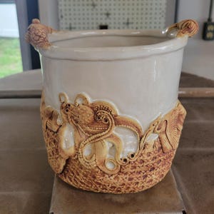 May include: A cylindrical ceramic vessel with a smooth off-white upper section and a textured, tan-coloured lower base. The exterior features relief carvings of an octopus and other marine life, with small sculpted fish figures on the rim.