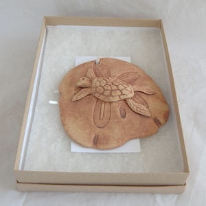 May include: A ceramic sand dollar ornament with a sculpted sea turtle design. The ornament is a light brown color and is displayed in a brown box.