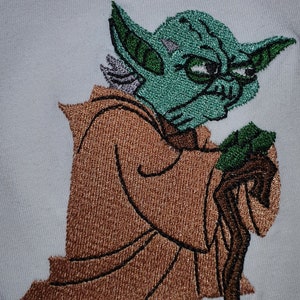 Peut inclure: Image brodée de Yoda de Star Wars, portant une robe brune et tenant un bâton de marche. Il a la peau verte, de grandes oreilles et une expression sérieuse.