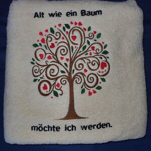 Może przedstawiać: Biały ręcznik z haftowanym wzorem drzewa. Drzewo ma brązowe gałęzie, zielone liście i czerwone serca. Tekst "Alt wie ein Baum möchte ich werden." jest wyszyty pod drzewem.