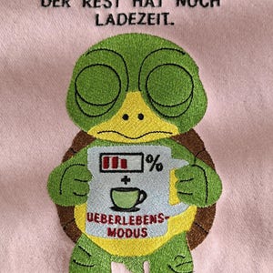 Puede incluir: Tela rosa bordada con un diseño de tortuga verde. La tortuga sostiene un letrero con un icono de batería, una taza de café y las palabras "UEBERLEBENS-MODUS". Encima de la tortuga está el texto "MEIN KÖRPER IST WACH. DER REST HAT NOCH LADEZEIT."