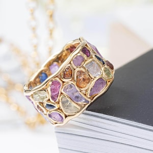 May include: A gold ring featuring a mosaic of colorful gemstones. The ring's design incorporates various shapes and hues, including pink, purple, orange, and blue stones, set within a gold band. The ring is displayed on a stack of white paper.