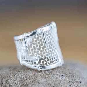 May include: A silver ring with a mesh design. The ring is wide and has a geometric shape.