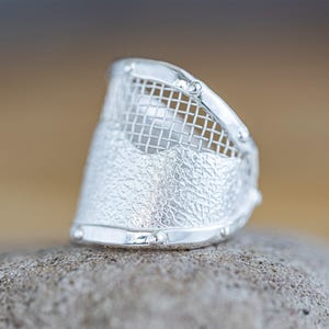 May include: A silver ring with a textured band and a mesh design on the top. The ring is sitting on a gray rock.