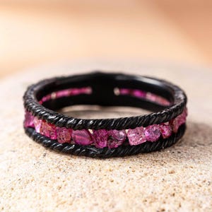 Puede incluir: Un anillo negro con un diseño de cuerda trenzada, con una fila de piedras preciosas rosas. Las piedras preciosas son de corte rugoso y añaden un toque de color a la banda oscura. El anillo está sobre una superficie de tono neutro.