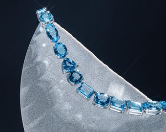 May include: A dazzling necklace featuring a series of light blue gemstones in various shapes, including oval, pear, and rectangular cuts. The gems are set in a silver or white metal, creating a striking contrast against the dark background.