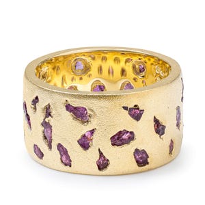 May include: A wide gold band ring with a textured matte finish, featuring multiple embedded raw, deep purple garnet stones scattered across the outer surface and visible along the inner circumference of the ring.
