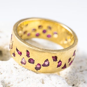 May include: A gold ring with a matte finish, featuring irregularly shaped openings filled with small, vibrant red gemstones. The ring is set against a white background, highlighting its design and color.