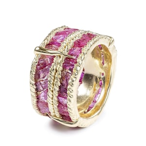 May include: A gold-colored ring featuring a band of vibrant pink gemstones. The ring has a textured, braided design around the gemstone band, with a small gold accent. The ring is set against a white background, highlighting its intricate details.
