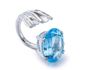 May include: A silver ring featuring a large, oval-cut, light blue gemstone. The ring's design includes a textured, leaf-like element and a band that curves around the finger, creating a unique and elegant look.