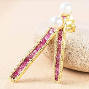 May include: Gold-toned earrings with a rectangular design, featuring a row of pink gemstones and a white pearl at the top. The earrings are set against a neutral background, showcasing their elegant design.