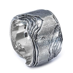 May include: A wide, textured silver ring with a unique, layered design. The ring has a rough, organic appearance, with visible details and a polished interior. The ring is marked with "925" indicating the silver content.