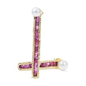 Peut inclure: Une paire de boucles d'oreilles dorées. Chaque boucle d'oreille présente une barre verticale de pierres précieuses roses, encadrée d'une bordure dorée torsadée. Une perle blanche est située en haut de chaque boucle d'oreille. Les boucles d'oreilles sont sur fond blanc.