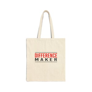 May include: A cream-colored tote bag with the words "DIFFERENCE MAKER" printed in red and black. The bag has two handles and is made of a sturdy fabric, suitable for carrying various items.
