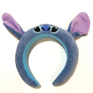May include: Blue plush headband with large ears and a stitched-on face of Stitch from Lilo & Stitch. The ears are purple and the face has black eyes and a blue nose.