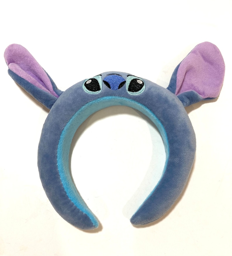 May include: Blue plush headband with large ears and a stitched-on face of Stitch from Lilo & Stitch. The ears are purple and the face has black eyes and a blue nose.