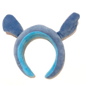 May include: Blue plush headband with large ears, resembling a cartoon character. The headband is made of soft, fuzzy fabric.