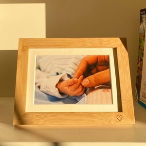 Può includere: Una cornice in legno con un passepartout bianco e un cuore intagliato nell'angolo in basso a destra. La cornice contiene una foto di due mani che si tengono.