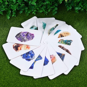 Könnte beinhalten: Eine Sammlung weißer rechteckiger Karten, die jeweils einen anderen farbigen Edelstein zeigen. Zu den Edelsteinen gehören Amethyst, Lapislazuli und andere Mineralien. Die Karten sind kreisförmig auf einer grünen Oberfläche angeordnet.