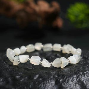 May include: A bracelet made of white, irregular gemstones, strung on a clear elastic cord. The bracelet is displayed on a dark, textured surface. The background is blurred, showing greenery and wood.