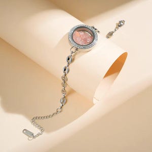 May include: A silver-toned bracelet featuring a round charm filled with pink gemstones. The charm is surrounded by a ring of small, clear stones. The bracelet has a delicate chain design with a lobster clasp.