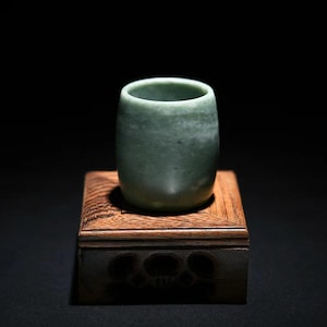 Könnte beinhalten: Eine kleine, zylindrische Jade-Tasse mit glatter, polierter Oberfläche. Die Tasse hat eine hellgrüne Farbe und steht auf einem quadratischen Holzsockel mit geschnitzten Details. Der Hintergrund ist schwarz.