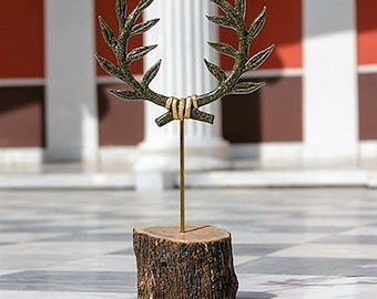 May include: A gold-colored olive branch wreath sculpture on a wooden base. The wreath is made of metal and has a green patina. The base is a natural wood slice.