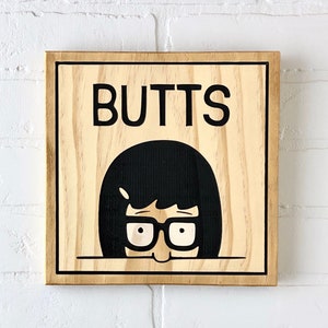May include: A wooden sign with the word "BUTTS" printed in black letters. A cartoon character with black hair and glasses is peeking out from behind the sign.