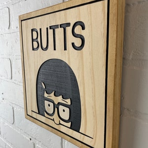 May include: A wooden sign with a black border and the word "BUTTS" in black lettering. A cartoon character with black hair and glasses is peeking out from behind the text.