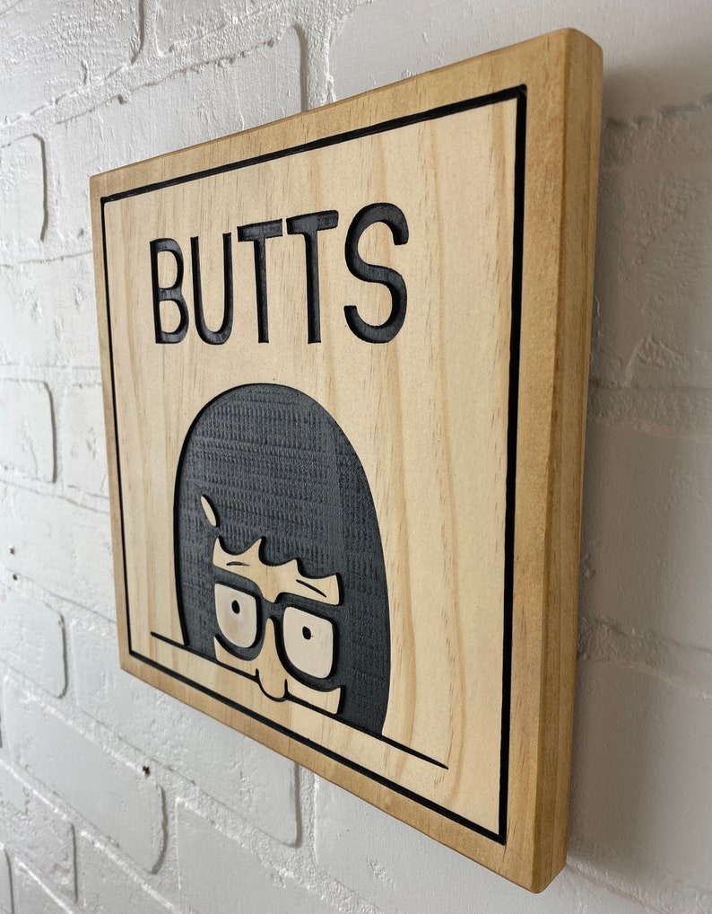 May include: A wooden sign with a black border and the word "BUTTS" in black lettering. A cartoon character with black hair and glasses is peeking out from behind the text.