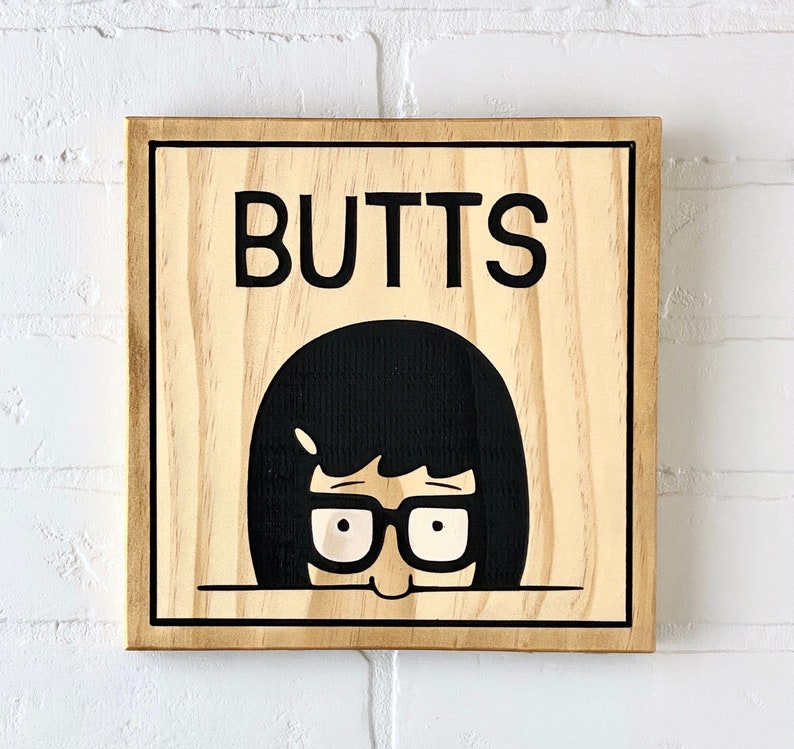May include: A wooden sign with the word "BUTTS" printed in black letters. A cartoon character with black hair and glasses is peeking out from behind the sign.