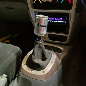 May include: An Asahi beer can serves as a gear shift knob in a car's interior. The car's dashboard is grey, with a digital clock showing 10:26. The gear shift is surrounded by a silver trim. The can is silver and white.