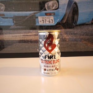 May include: A silver can of FIRE EXTREME BLEND coffee with a red flame logo. The can has a geometric pattern and Japanese text. The background features a blue car image.