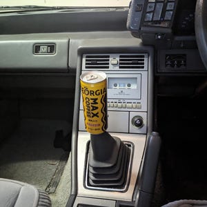 May include: Interior view of a classic car's console. A yellow and black "Georgia Max Coffee" can serves as a gear shift knob. The console includes a cassette player, air vents, and several buttons.