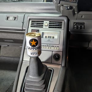 Pode incluir: Uma lata de cerveja Sapporo com um logotipo de estrela dourada está equilibrada no topo da alavanca de câmbio de um carro. O interior do carro é cinza, com um toca-fitas e saídas de ar visíveis no painel.