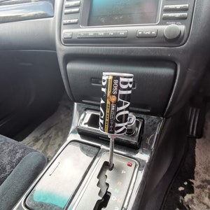 May include: Interior shot of a car's console featuring a custom gear shift. The shift lever is a can of "BOSS BLACK" coffee. The console is black with silver accents, and the car's dashboard is visible in the background.