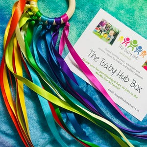 May include: A wooden ring toy with a rainbow of satin ribbons. The ribbons are attached to the ring and cascade downwards. A card with the text "The Baby Hub Box" is also in the image.